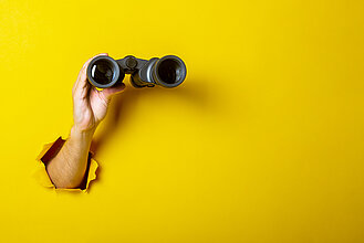 Female hand holds black binoculars on a yellow background. Looking through binoculars, journey, find and search concept.